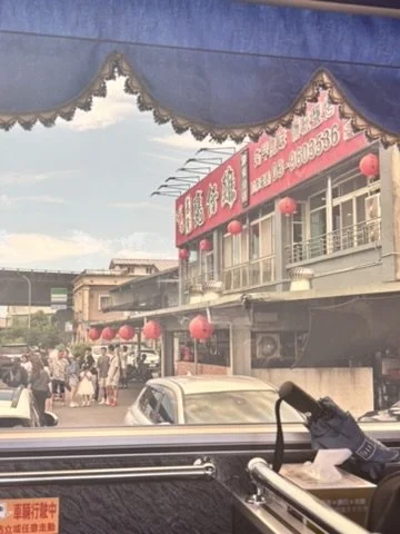 Street scene viewed from inside a vehicle with a window display showing a building with red signage and red lanterns, people walking on the sidewalk, cars parked on the street, and a blue canopy above.