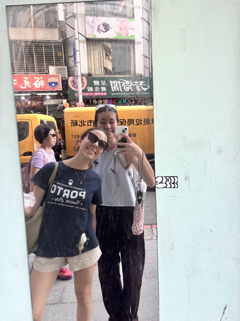 Two young women taking a selfie in a mirror on a city street. One wears sunglasses, a black t-shirt, and beige shorts; the other wears a striped shirt and has a backpack. Behind them are pedestrians, a yellow truck, and storefronts with signs.