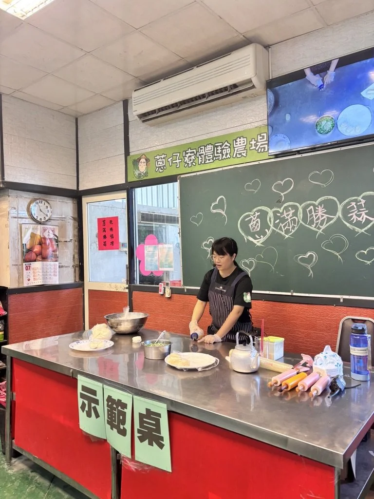 A woman standing behind a stainless steel counter at an indoor demonstration table, with kitchen tools, ingredients, and rolled cookies in front of her. There is a wall with a blackboard decorated with hearts and Chinese writing, a wall-mounted air c
