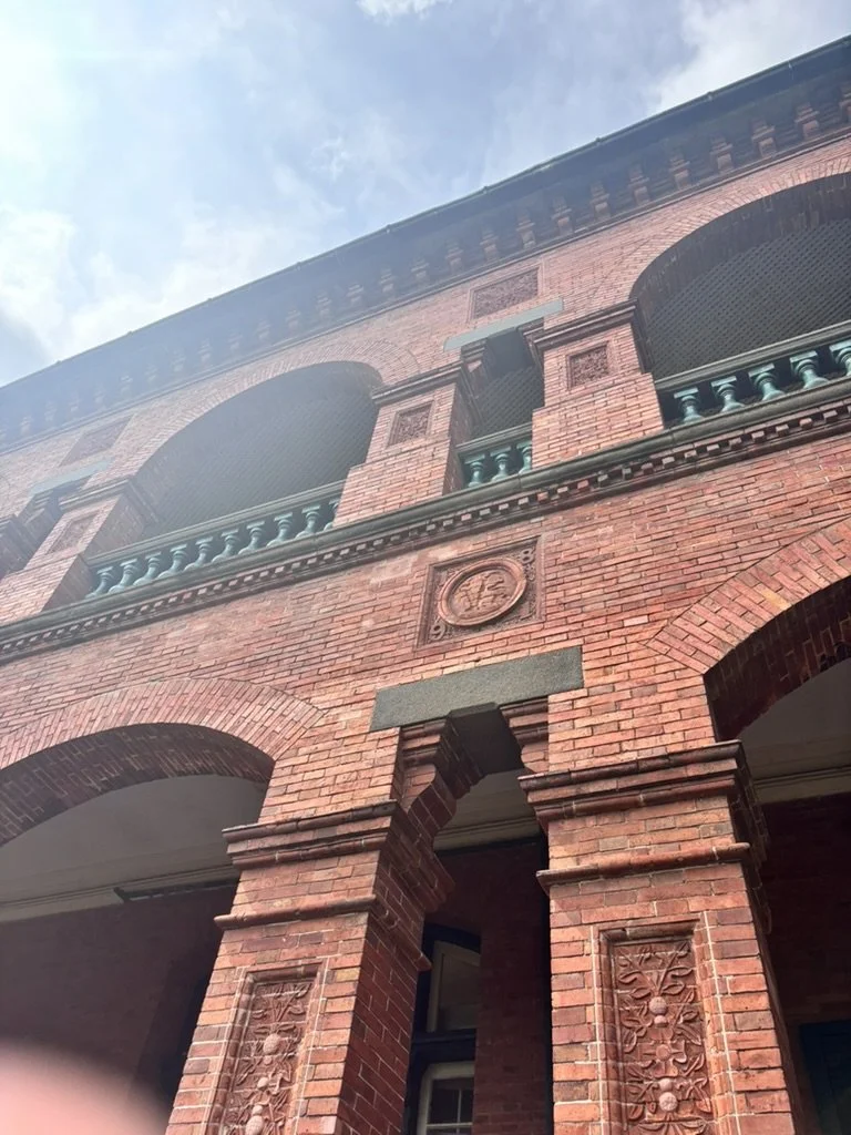A red brick building with arched windows, decorative stone carvings, and a small balcony with teal railing, seen from below against a blue sky with some clouds.