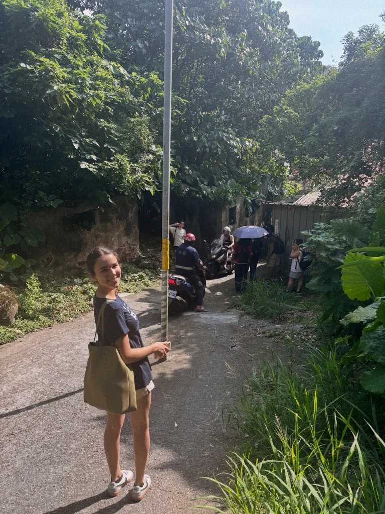 A young girl holding a pole on a narrow, shaded path in a green, outdoor area. In the background, there are police officers, a person with an umbrella, and some people near motorcycles and a small building. The scene appears to be in a rural resident