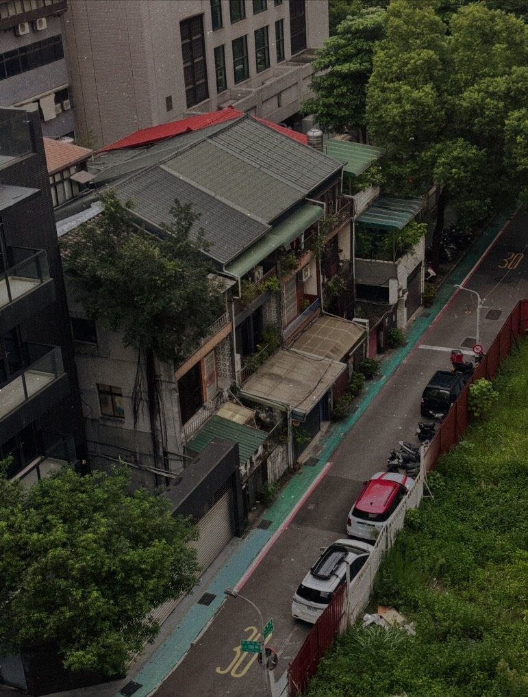 A view from above of a narrow urban street with parked cars and motorcycles, alongside small residential buildings with metal roofs and green awnings, bordered by tall modern buildings and greenery.