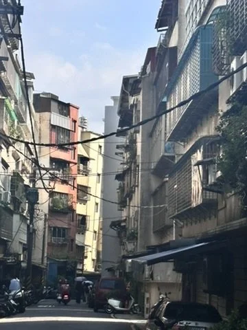 Narrow city street lined with multi-story buildings, some with balconies and air conditioning units, with cars and motorbikes parked along the sides.