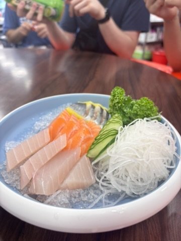 A bowl of sashimi with slices of salmon and tuna, garnished with cucumber slices, broccoli, shredded daikon, and wasabi, on a dark wooden table.
