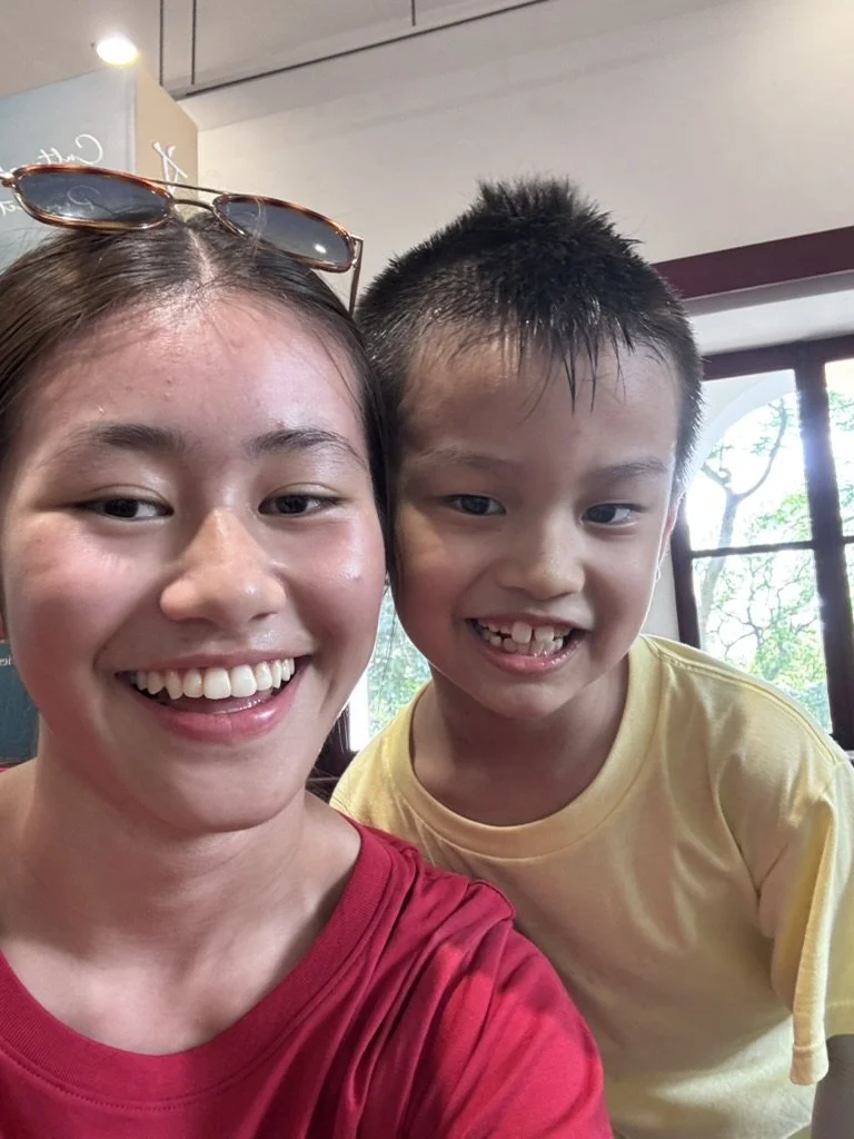 A woman and a boy smiling for a selfie indoors, with a window showing trees outside.