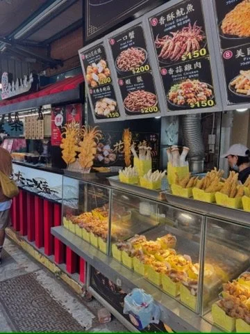 Asian food stall with menu boards displaying various dishes and prices, and a glass display case filled with fried food items like chicken pieces, along with yellow containers for utensils or napkins.
