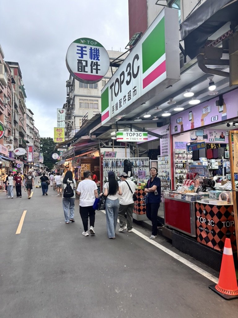 Busy street market with various shops, people browsing and walking, signs in English and Chinese, and overcast sky.