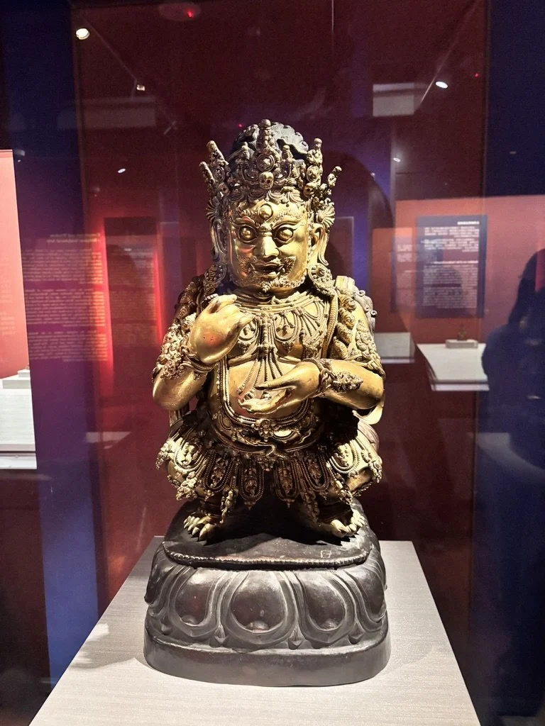 A detailed gold-colored statue of a mythological figure with multiple heads, intricate jewelry, and elaborate clothing, displayed in a museum case with red-colored background panels.