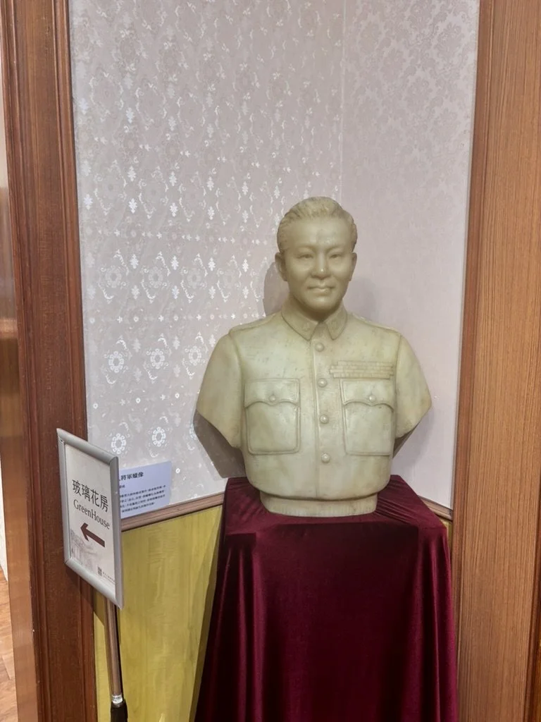 A bust of a woman in military uniform displayed on a pedestal with a red cloth, situated in a room with textured wallpaper and wooden paneling, and a sign indicating the direction to a greenhouse.
