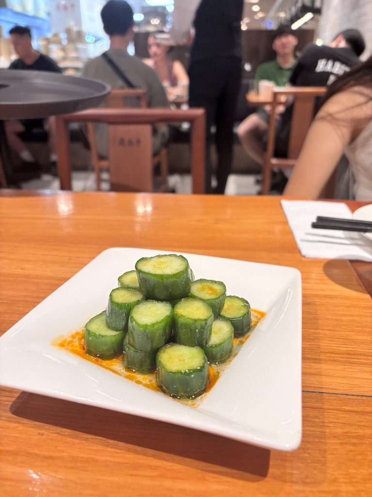 A plate of sliced cucumber rolls arranged in a pyramid on a white square dish, with a reddish sauce around the base, on a wooden table in a restaurant.