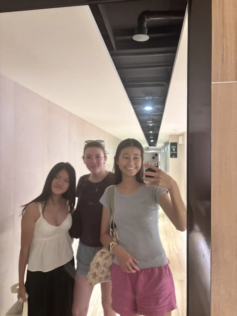 Three young women taking a mirror selfie in a hallway, smiling at the camera.