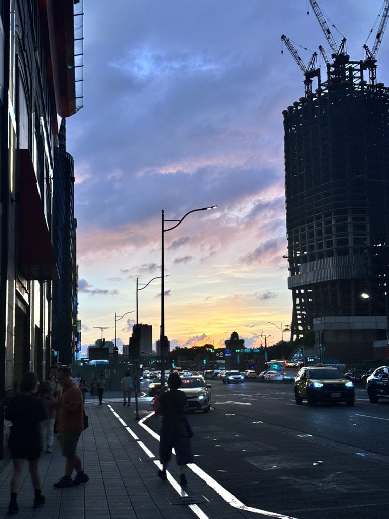City street at sunset with a mix of pedestrians and traffic, tall buildings, and construction cranes in the background.