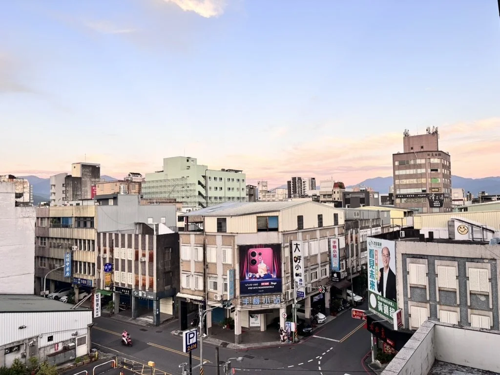 Cityscape of a small urban area during sunset with buildings, billboards, and a street with vehicles.