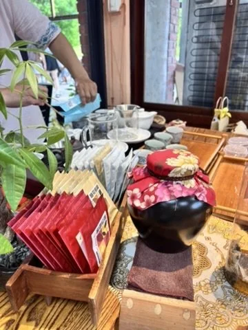 A table set with various dishes, cups, teapots, and a large decorative pottery jar covered with a cloth featuring floral patterns. Red packets are arranged in a wooden box, and a person is seen in the background near a window.