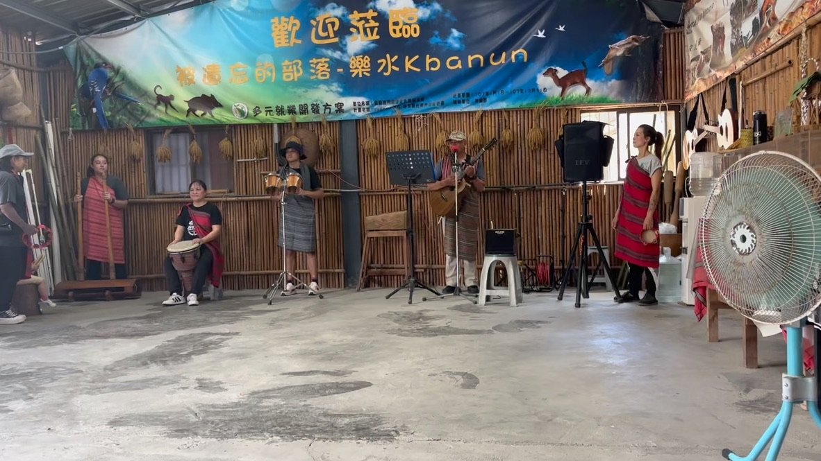 Group of musicians performing in a bamboo-walled indoor space, with a large colorful banner overhead and various musical instruments. A woman is seated with a drum, two women and one man are playing guitars, and two women are standing nearby. A large