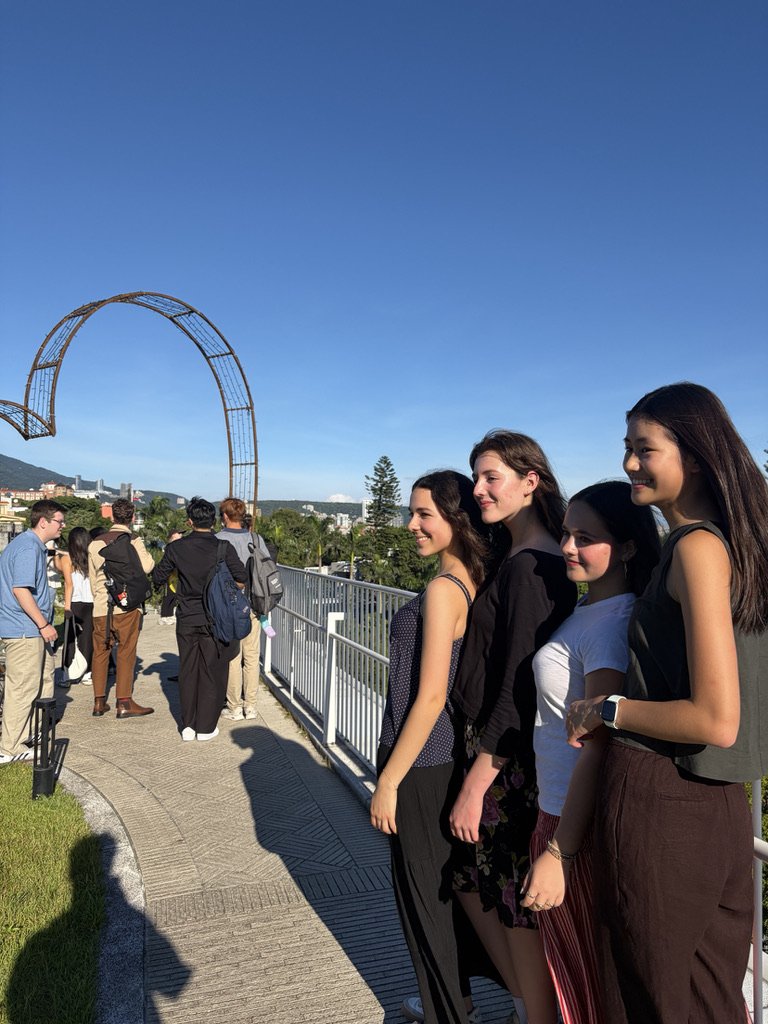 A group of four young women standing on an outdoor walkway, smiling and posing for a photo with a city skyline and a large, curved metal structure in the background on a sunny day.