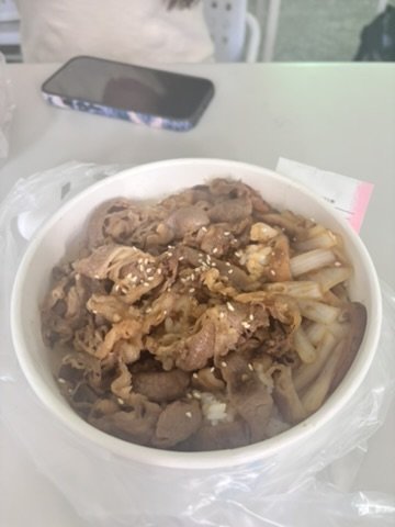 A bowl of Asian noodle soup with sliced meat, possibly beef or pork, topped with crushed garlic or sesame seeds, on a white table with a smartphone in the background.