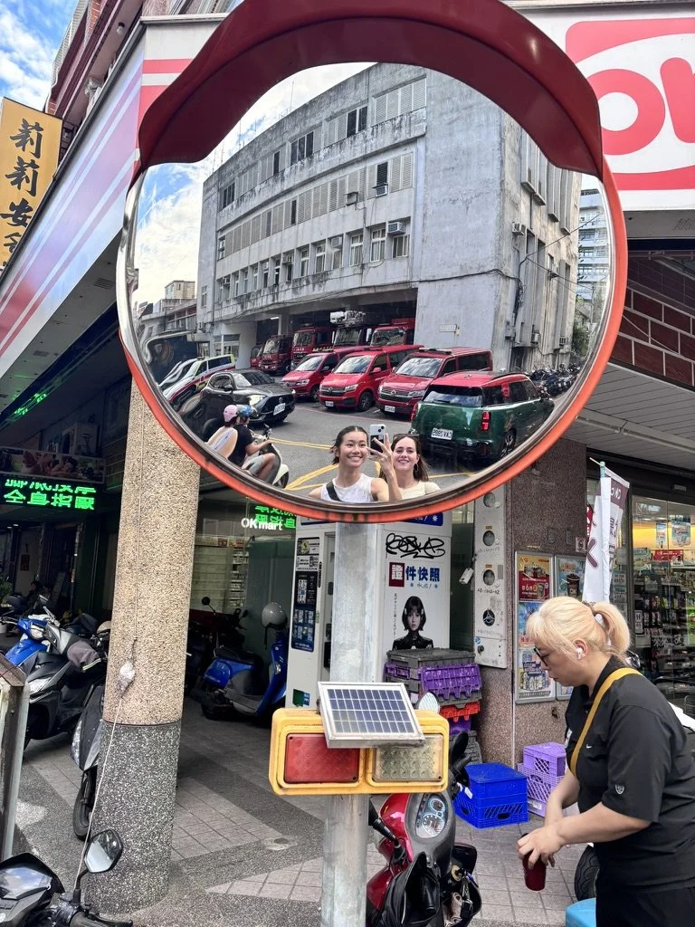 A street scene featuring a convex traffic mirror reflecting two smiling women taking a selfie, parked cars and scooters, and a building with multiple windows. In front, a fair-skinned woman with blonde hair tied back, wearing a black shirt and carryi