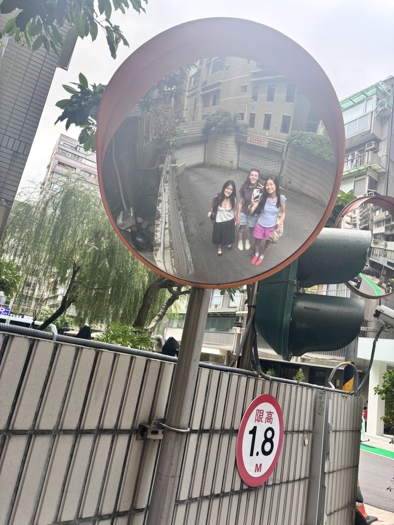 Three people taking a selfie in a convex street mirror in an urban area, with buildings and trees reflected in the mirror.