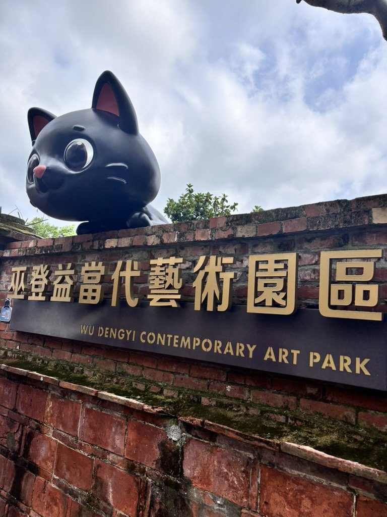 Large black cat statue with big eyes and pink nose on brick sign at Wu Dengyi Contemporary Art Park, with cloudy sky in background.