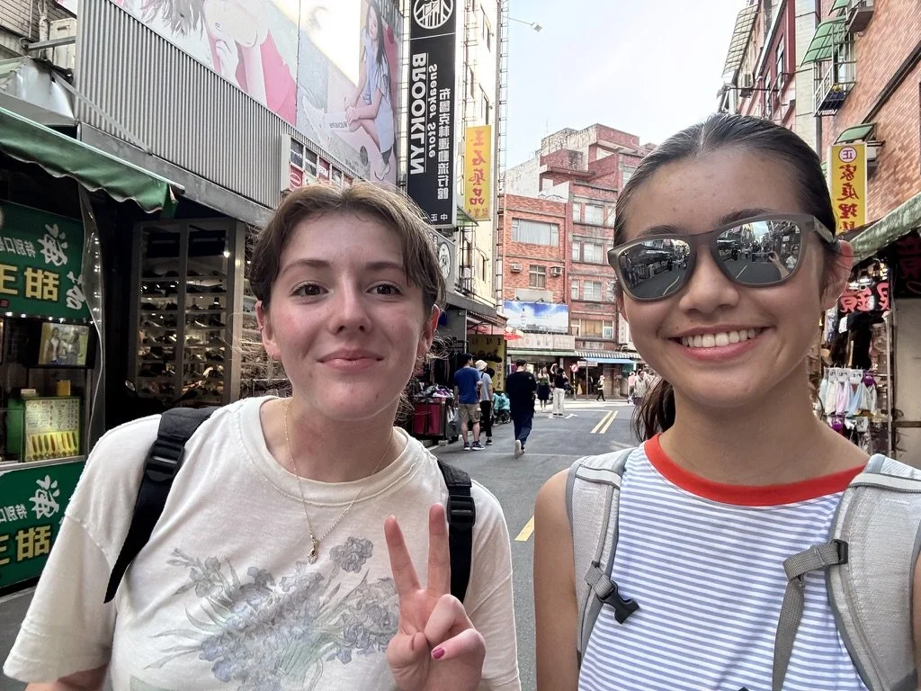 Two young women smiling and taking a selfie on a busy city street in an Asian country, with buildings, shops, and signs in the background.