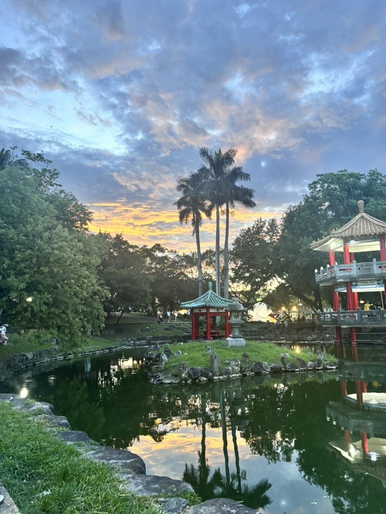 A serene park scene during sunset with a pond, traditional Asian-style pavilion, red lanterns, and palm trees reflected in the water.