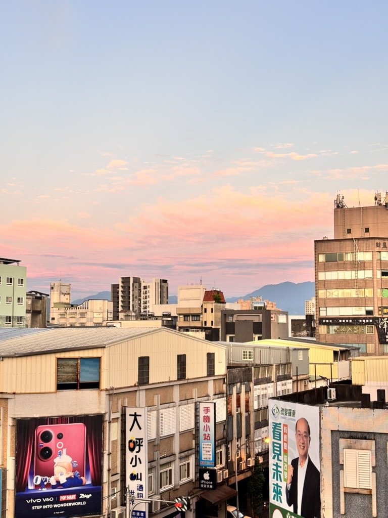 Cityscape of buildings with colorful sunset sky and mountains in the background, advertisements on buildings, including a pink smartphone banner and a man in a suit with glasses.