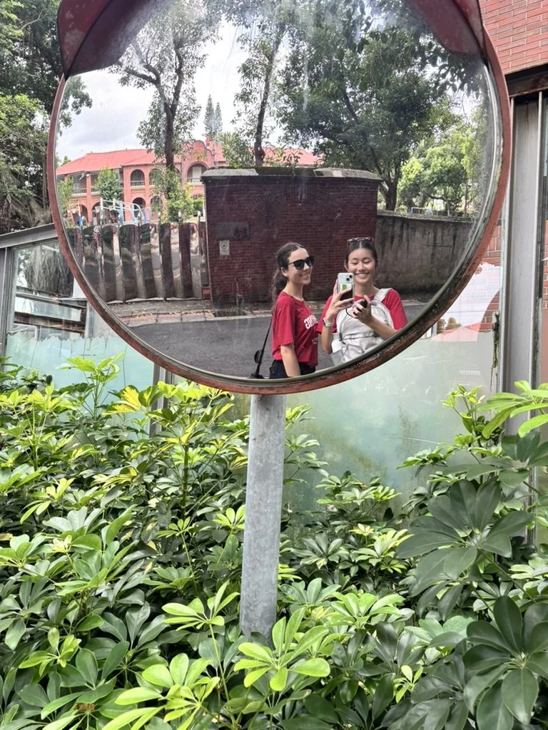 Two women taking a selfie in a round mirror reflection, with greenery and trees in the background.