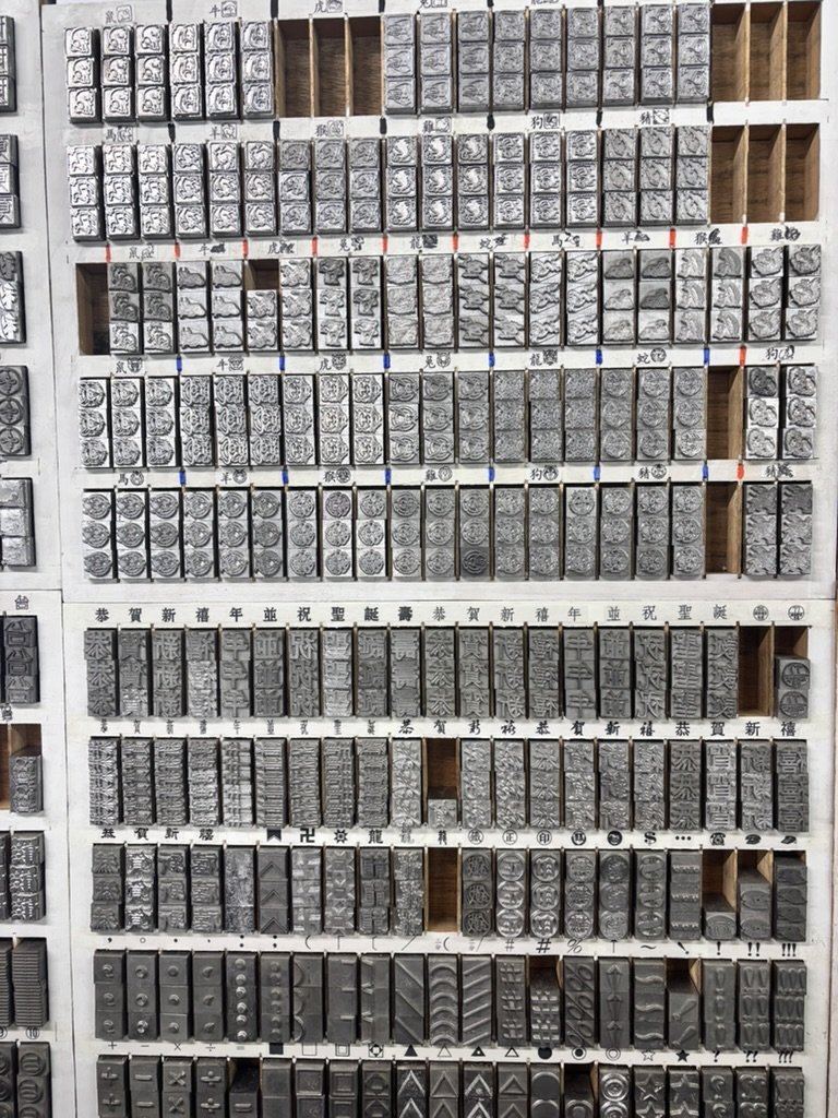 Display of various metallic rubber stamps with Chinese characters, symbols, and patterns organized in rows on a white wooden shelf.