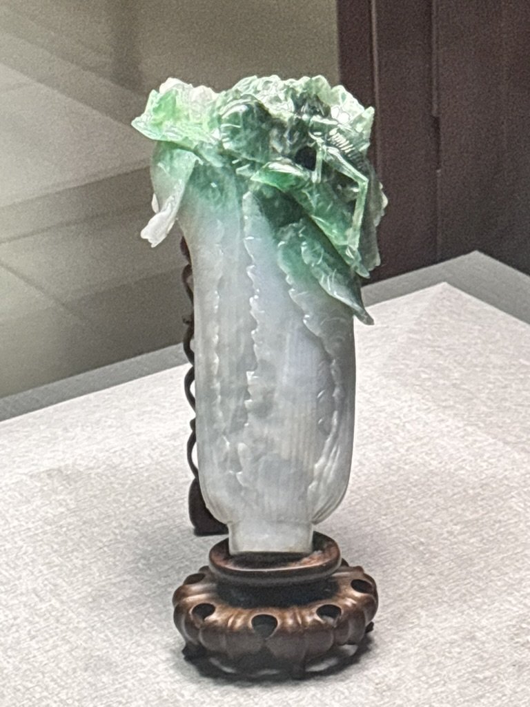A decorative jade or onyx sculpture resembling a vase with green leafy carvings at the top, mounted on a carved wooden base.