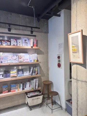 Bookshelves with books and magazines, a small cooler, a stool, and two chairs in a room with concrete walls and ceiling.