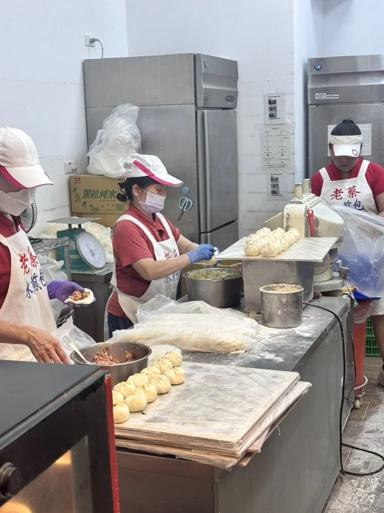 Three women working in a kitchen preparing steamed buns. They are wearing hats, masks, and gloves, and using various tools and ingredients for baking.