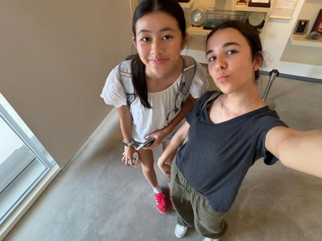 Two young girls taking a selfie inside a building, with display cases and shelves in the background.