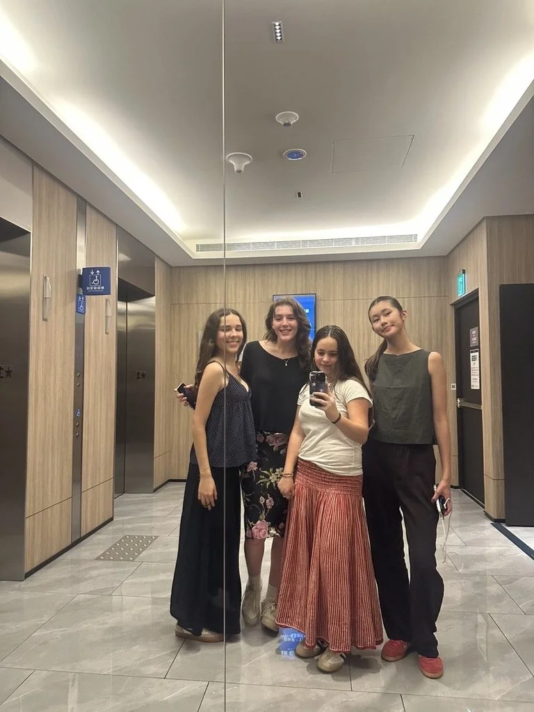 Four young women standing in front of a mirror, taking a selfie in a modern building lobby with wood-paneled walls and elevator doors.