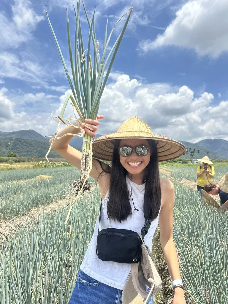 Smiling woman in sunglasses and a wide-brimmed straw hat holding a large onion or garlic plant in a field of green crops, with mountains and a partly cloudy sky in the background.