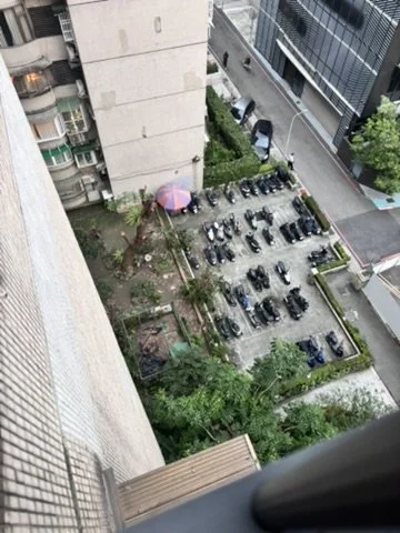 View of a city parking lot with cars and motorcycles from a high vantage point, next to multi-story buildings and a street with some greenery.