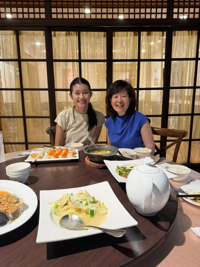 Two women sitting at a dinner table with various dishes, smiling at the camera in a restaurant with wooden decor.
