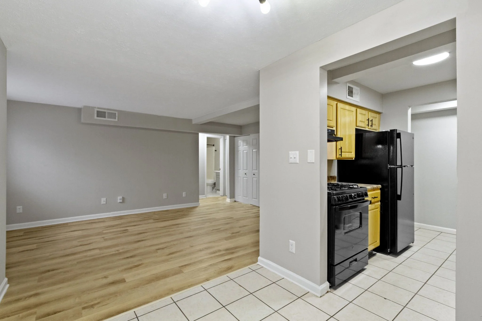Open-concept living area with beige walls, light wood flooring, and a kitchen with yellow cabinets, a black refrigerator, and black stove, and a doorway leading to a bathroom.