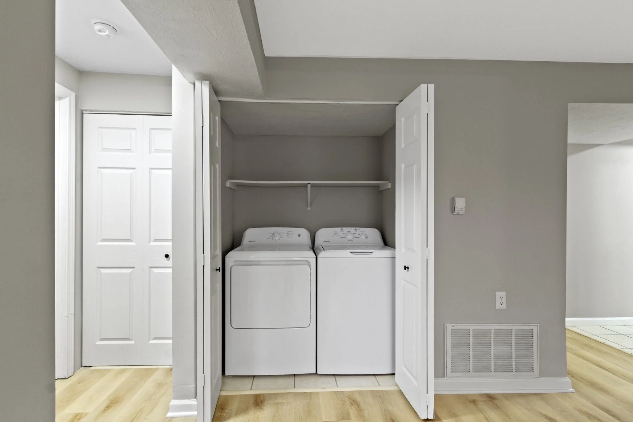 A laundry area with a white washer and dryer in a closet with open white bi-fold doors, gray walls, and light wood flooring.