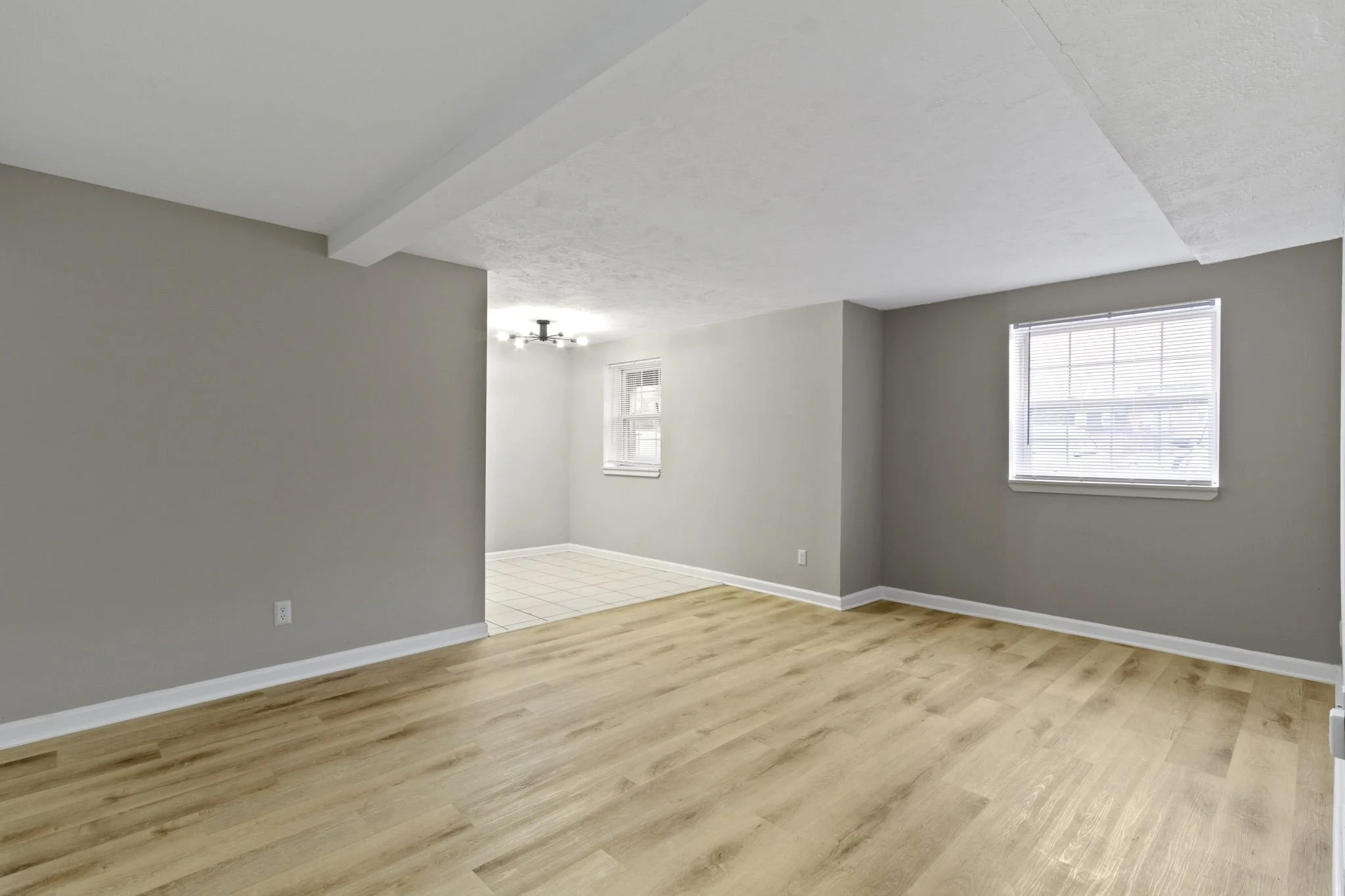 Empty living room with wood flooring and windows, connected to a dining area with tile flooring and a ceiling light fixture.
