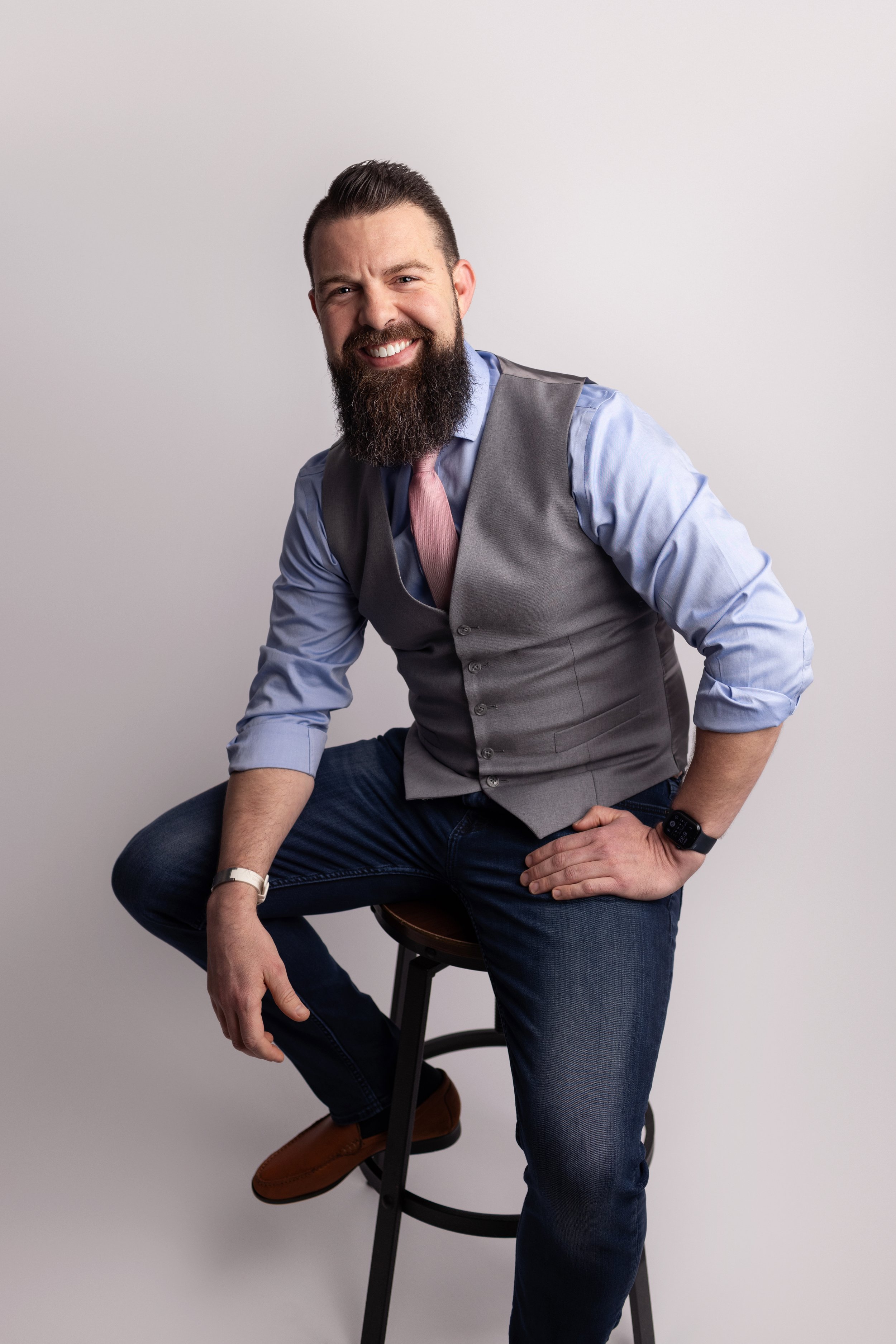 A man with a beard and short styled hair smiling while sitting on a stool against a plain gray wall. He is wearing a light blue shirt with rolled-up sleeves, a gray vest, and dark jeans. He has a smartwatch on his left wrist and a pink tie, with his right hand resting on his knee and his left hand on his thigh.