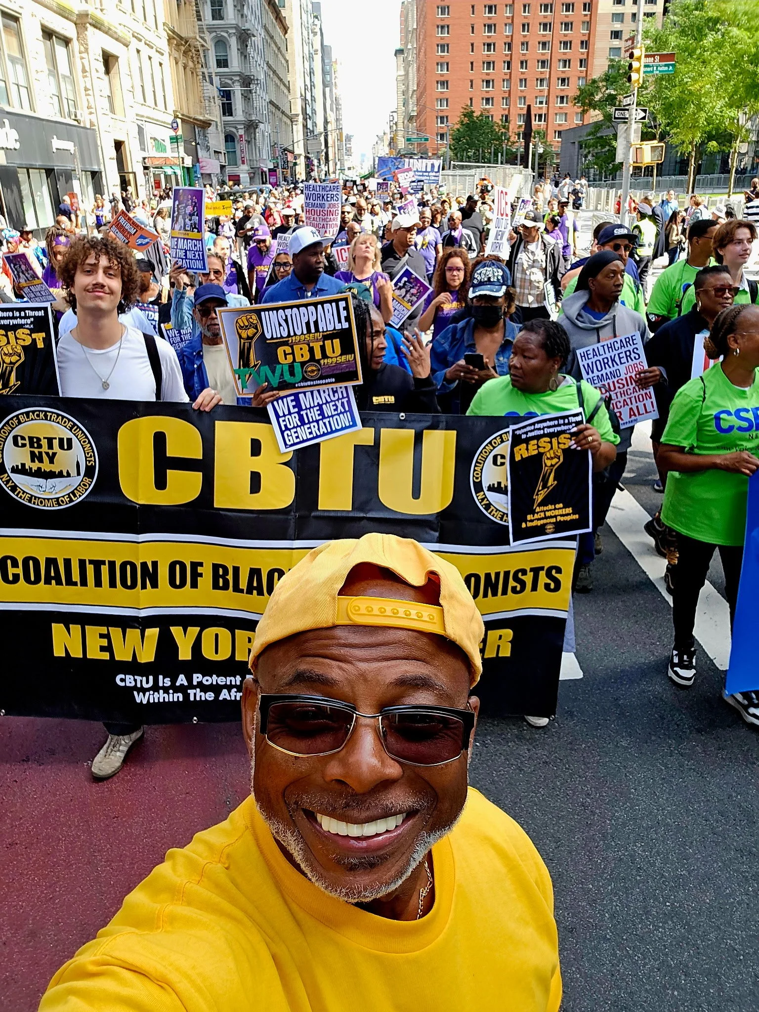 A man wearing sunglasses, a yellow cap, and a yellow shirt taking a selfie at a protest march. Behind him, a large crowd of people holding signs and banners advocating for workers' rights and social justice, with city buildings and trees in the background.