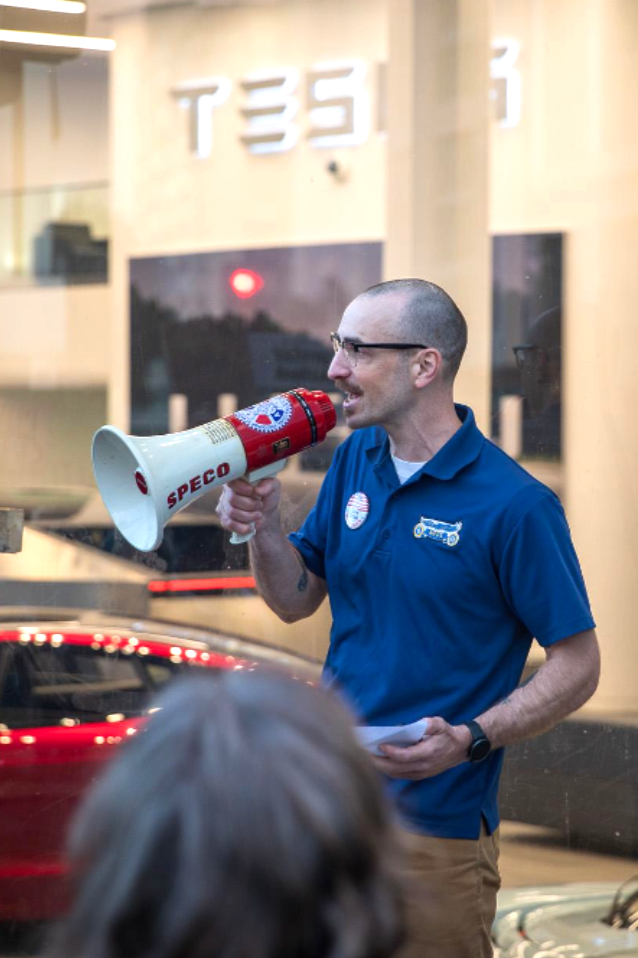 A man with glasses, bald head, wearing a blue polo shirt with patches, holding a megaphone and speaking to a crowd inside a building at sunset.