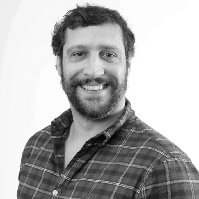 Smiling man (CEO Nuno) with brown hair and beard, wearing a dark plaid shirt, against a white background.