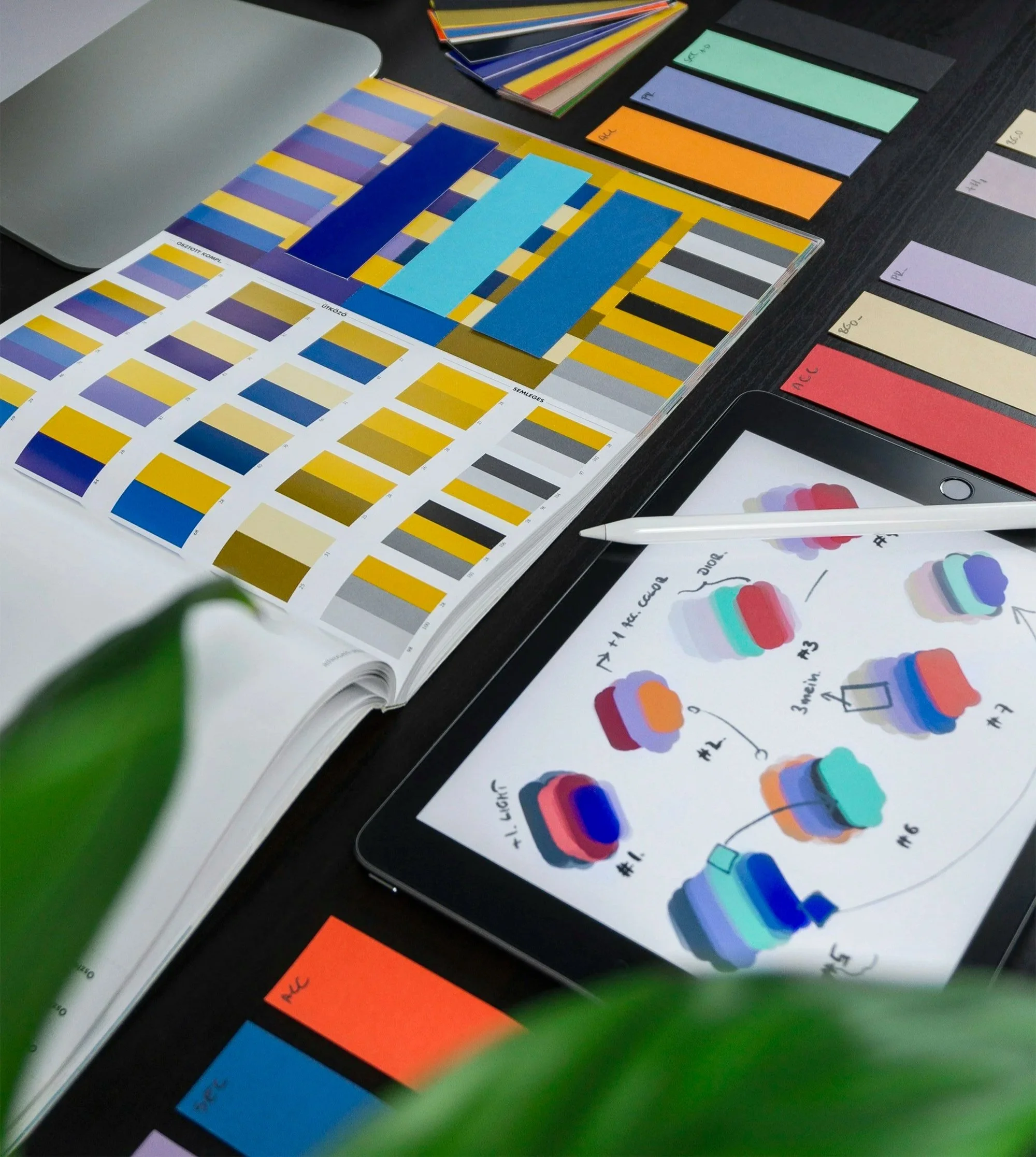 Work desk with color samples and palettes, a digital tablet displaying color diagrams, and a fountain pen alongside paper filled with sketches and notes exploring color combinations.