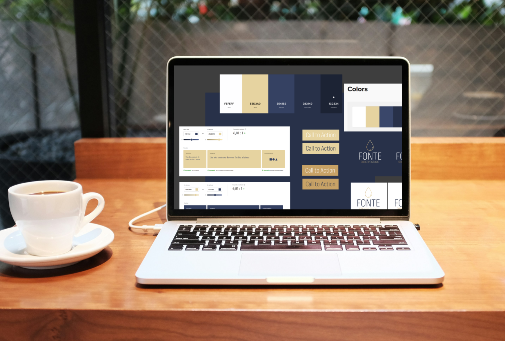 Laptop on a wooden desk displaying a color design and call-to-action buttons, with a coffee cup beside it, set in front of a window with a wire fence in the background.