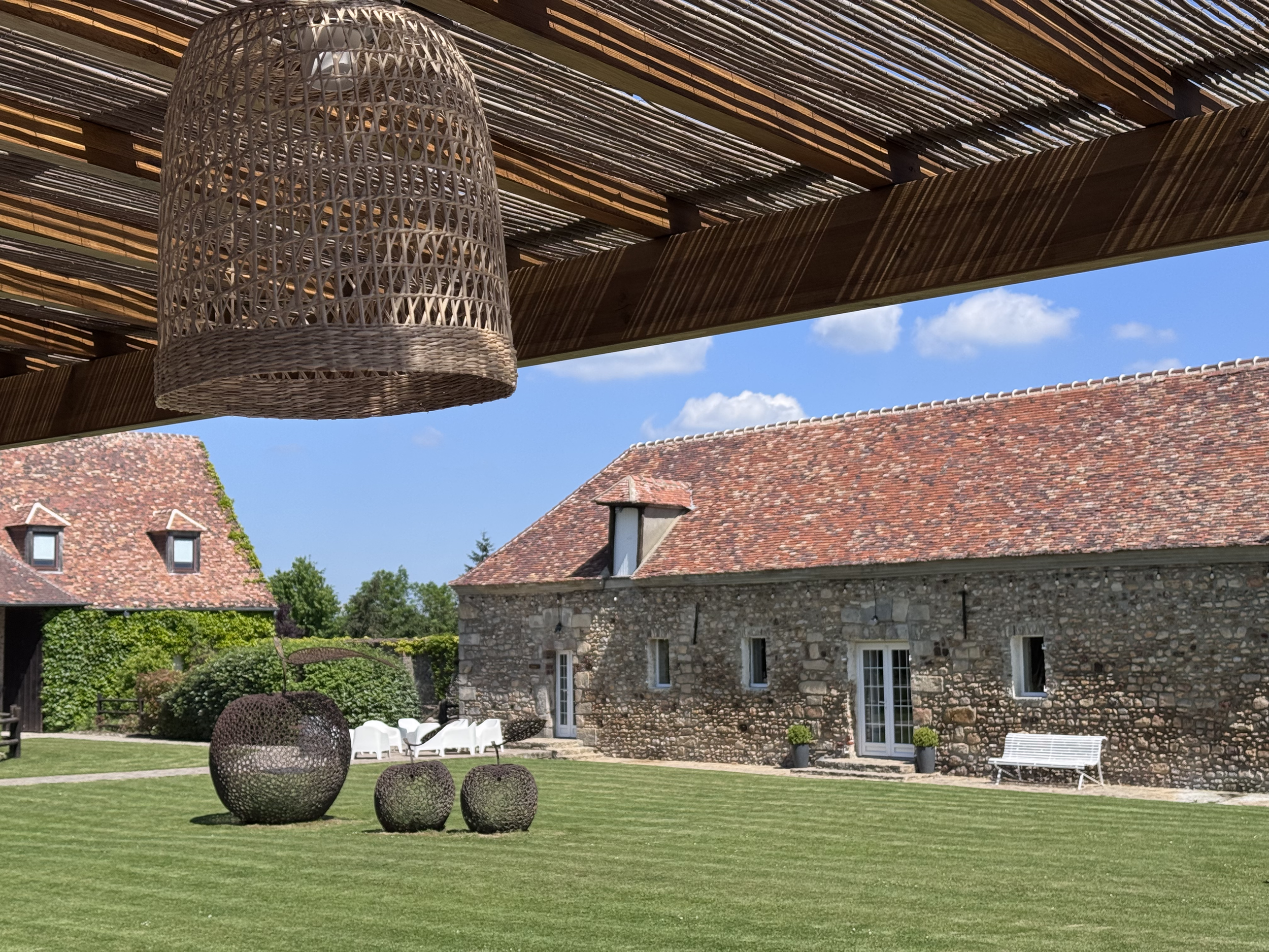 Une cour extérieure avec un bâtiment en pierre, des meubles en rotin noir, une pelouse verte bien entretenue, et un ciel bleu avec quelques nuages. Un lampadaire en rotin suspendu est visible sous un auvent en bois et bambou.