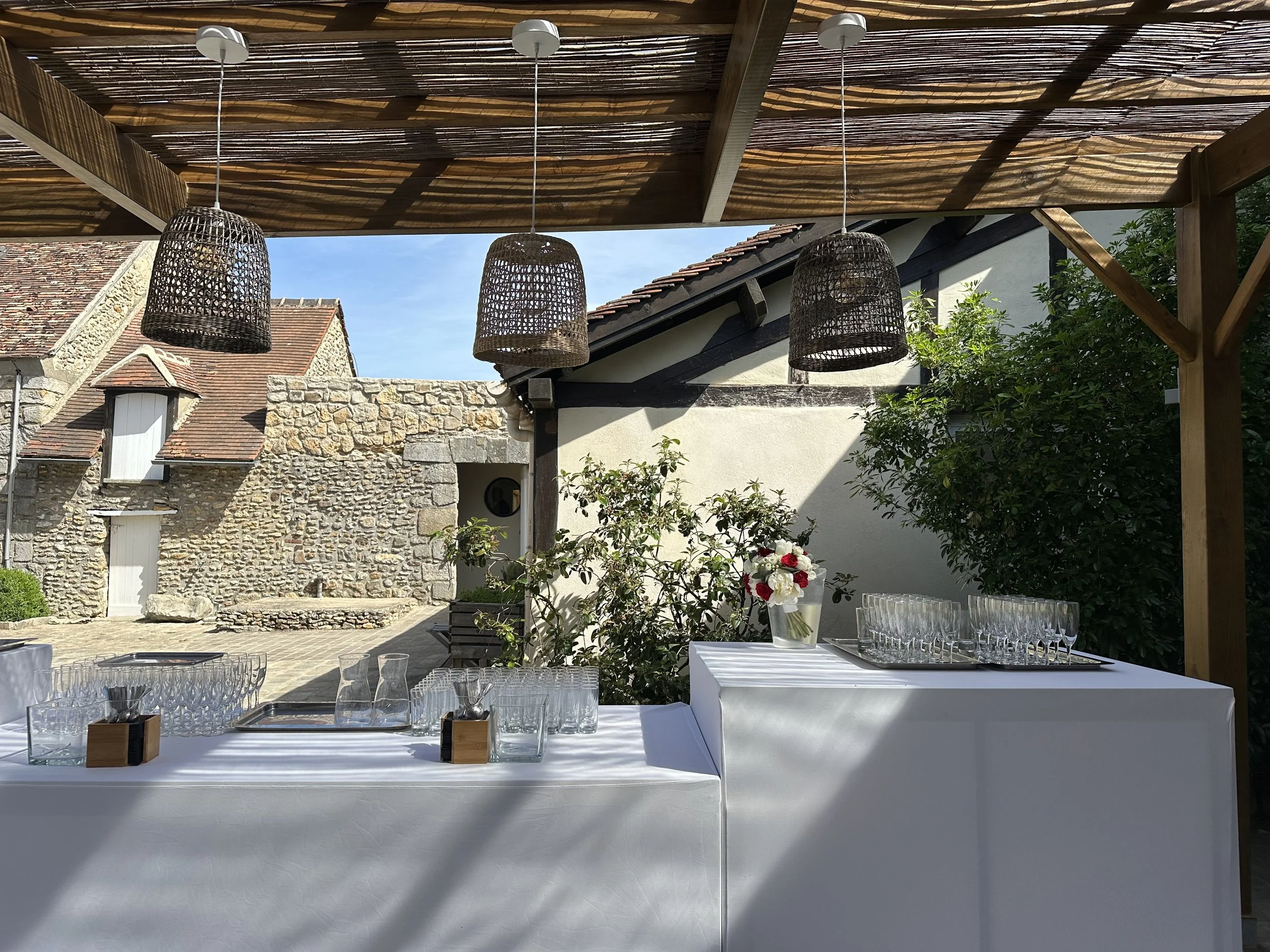 Une table blanche décorée pour un repas en extérieur sous un auvent en bois avec des lampes en rotin suspendues.