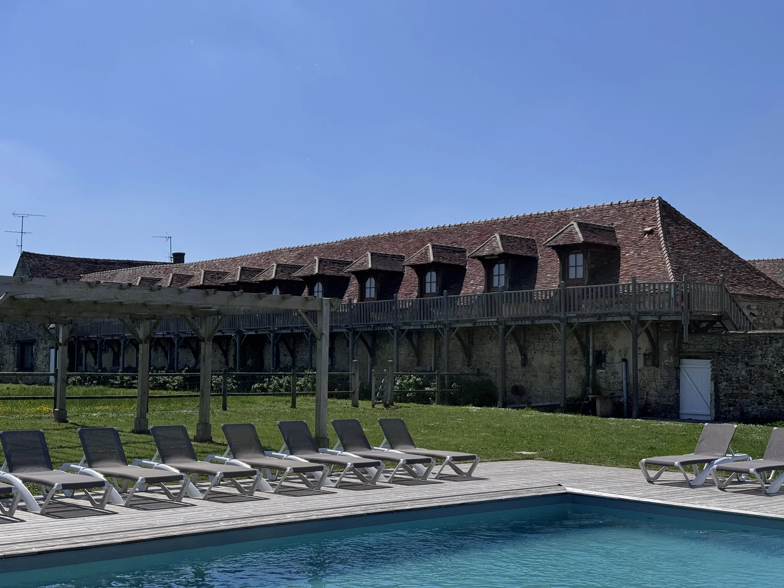 Piscine extérieure avec chaises longues en plastique blanc, rivière ou bassin au premier plan, bâtiment en pierre avec toit en tuiles rouges et fenêtres en dormer en arrière-plan, ciel clair et bleu