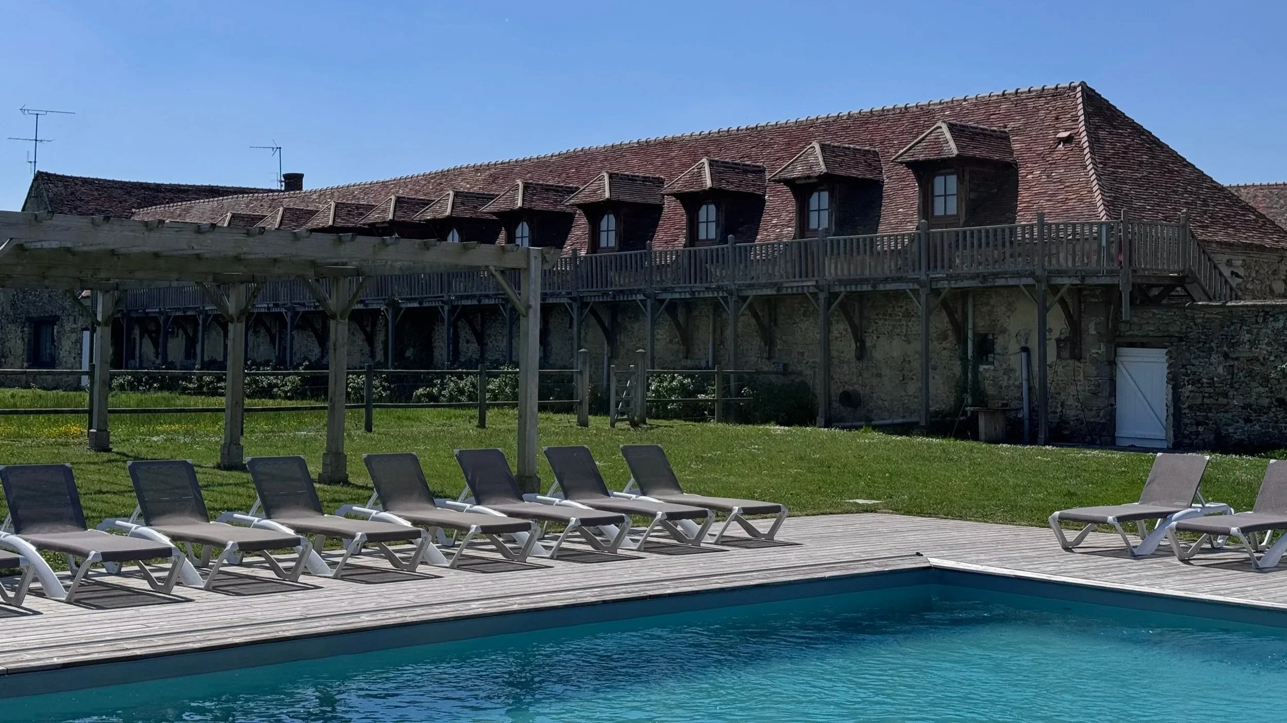 Piscine extérieure avec des chaises longues en plastique blanc sur un pont en bois, une maison en pierre avec un toit en tuiles rouges et une terrasse en bois, et un ciel clair